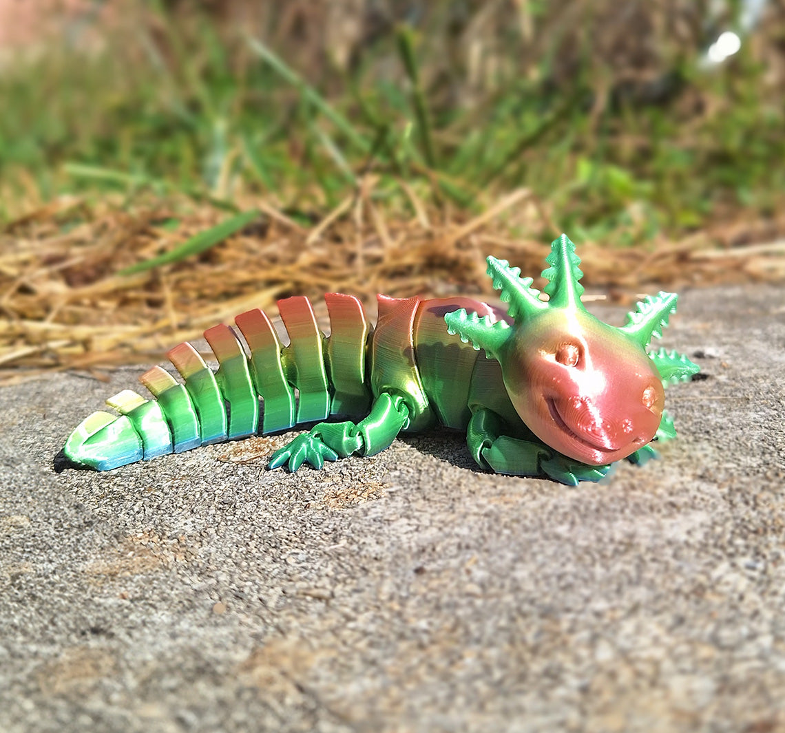 A flexible, articulated axolotl figure resting on textured concrete, crafted from Eryone Rainbow Candy Silk PLA filament that boasts a gradient of red to green. The figure's design allows for sensory interaction, providing a tactile fidget experience as each segment moves. The background blurs into a natural setting, highlighting the figure as a captivating piece for sensory stimulation and mindful fidgeting.
