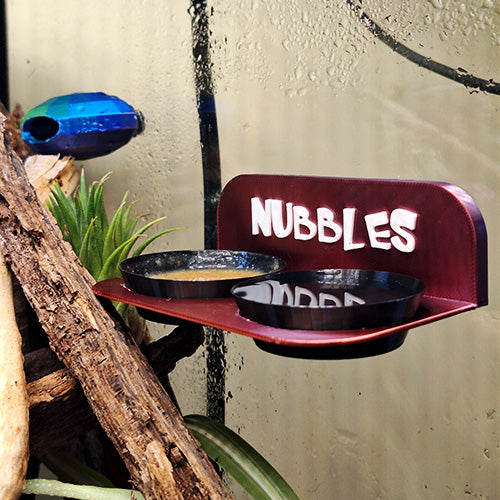 Magnetic Gecko Feeding Ledge with customizable cups, holding crested gecko diet on the left and water on the right. A blue mini gecko hide is positioned in the top left corner for added enrichment and habitat variety.