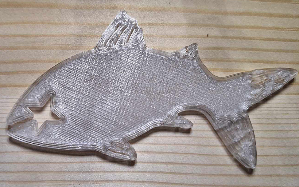 Overture translucent PETG Fishy Bite Bread & Bag clip showcasing its clear, shimmering quality on a wooden surface. The fish silhouette clip is both practical and aesthetically pleasing, with a transparent finish that allows the texture of the wood grain to show through.