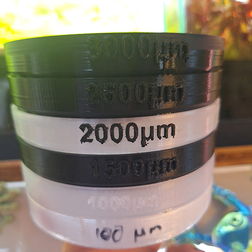 Stack of 3D-printed mesh sieves with varying micron sizes labeled on each layer, used for precise filtering, against an illuminated backdrop.