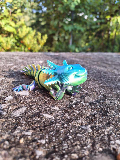 An articulated axolotl toy posed on a rough concrete surface, 3D-printed in Aceaddity Blue/Yellow/Purple tri-color silk PLA filament, with color transitions creating a vibrant, lifelike appearance. The toy is set against a natural outdoor backdrop, its intricate details and bright colors standing out, making it a captivating piece for collectors and educational play.