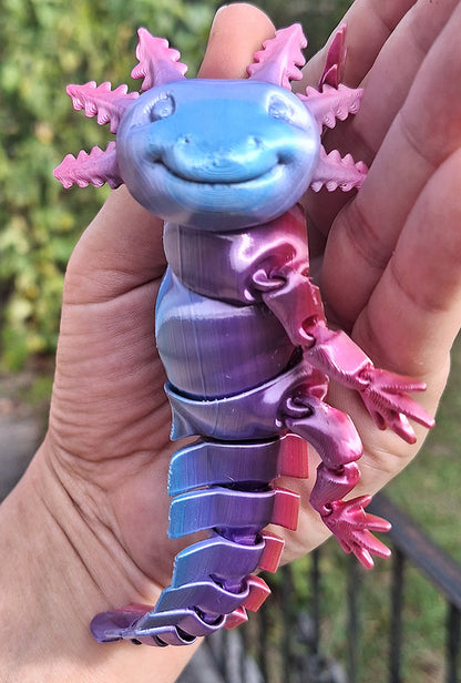 A hand displaying an articulated axolotl figure printed with Eryone Rainbow Candy Silk PLA filament, showcasing a playful smile and a spectrum of purple and pink hues. The figure's segments are clearly defined, allowing for movement, and it's set against a blurred garden backdrop, illustrating a cheerful and educational item perfect for enthusiasts of creative 3D printing.