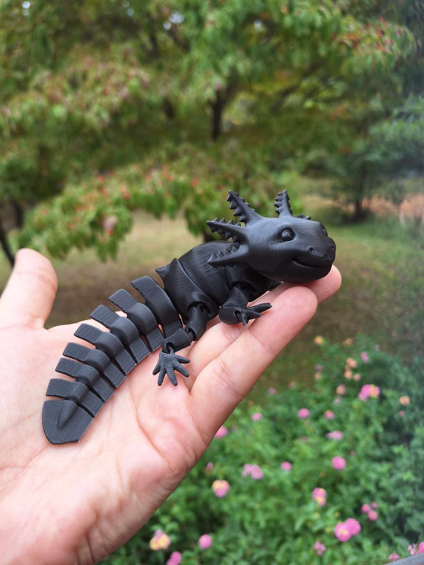 A hand presents an articulated axolotl figure printed in Creality Hyper Black PLA filament, offering a sleek, matte finish. The figure is designed for sensory engagement, with segments that provide a satisfying fidget experience. Positioned against a garden with blurred foliage and flowers, the piece serves as a tactile object for sensory exploration and stress relief.