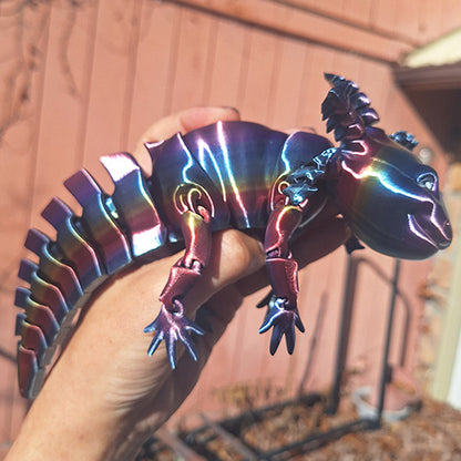 A hand displays a multi-colored articulated axolotl figure, shimmering with a metallic rainbow sheen, against a rustic backdrop.