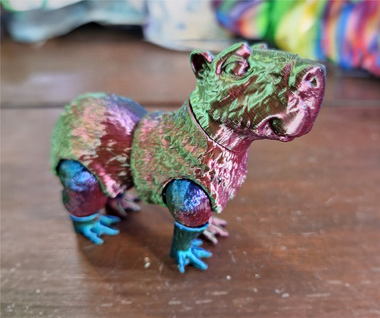 An articulated capybara figure printed in Eryone Silk PLA, featuring a lustrous color blend of red, blue, and green. The figure captures the capybara's gentle expression and relaxed posture, with each segment designed to move, providing a tactile experience. Placed on a wooden surface, the figure's shimmering silk finish and vibrant colors make it a visually striking piece for interactive display and sensory exploration.