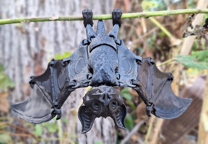 An articulated fruit bat figure hangs upside down from a twig, 3D-printed in Creality Hyper Black PLA, with detailed wings outstretched. The figure is displayed against a natural backdrop, with the texture of the filament providing a realistic look to the model. The bat's design allows for movement in the wings, presenting a natural pose that blends seamlessly with the outdoor environment.