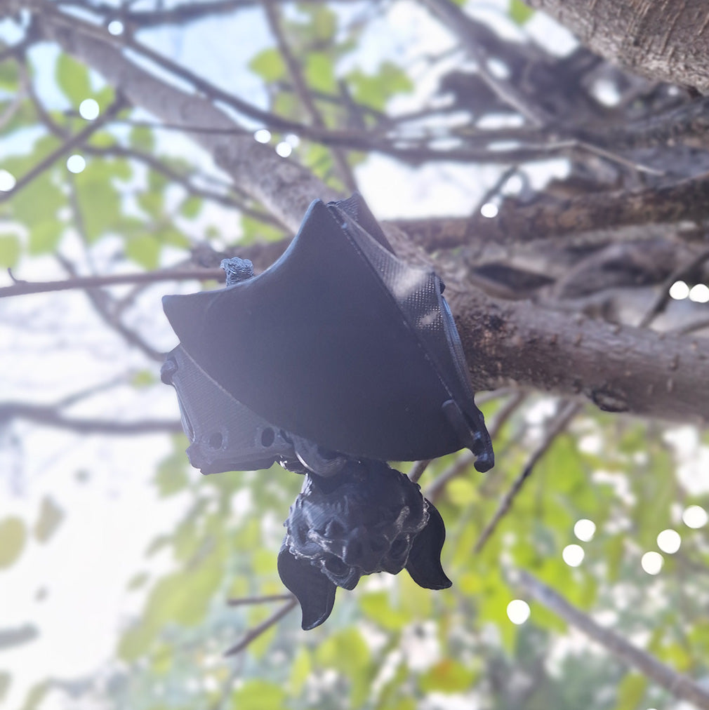 3D-printed black Articulated Fruit Bat in an upside-down pose with its wings wrapped around its body, attached to a tree branch, with a blurred background of green leaves highlighting the bat's intricate design and flexibility, crafted from durable Creality Hyper PLA.