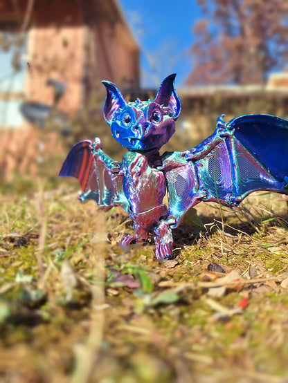 An articulated fruit bat figure stands on the ground, crafted from Aceaddity Tri-Color Silk PLA with vibrant red, blue, and green hues. The bat's wings are spread wide, demonstrating the flexible joints and the shimmering silk finish of the material. Set in a natural outdoor scene, the figure's colors are vivid against the earthy background, offering a visually stimulating and tactile experience for sensory play and fidgeting.