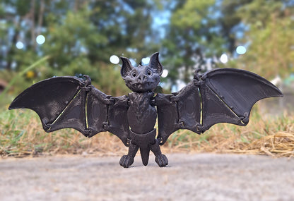 Black Creality Hyper PLA 3D-printed Articulated Fruit Bat standing with wings fully spread, centered on a concrete surface with a soft-focus background of trees, demonstrating the model's stability and detailed wing texture.