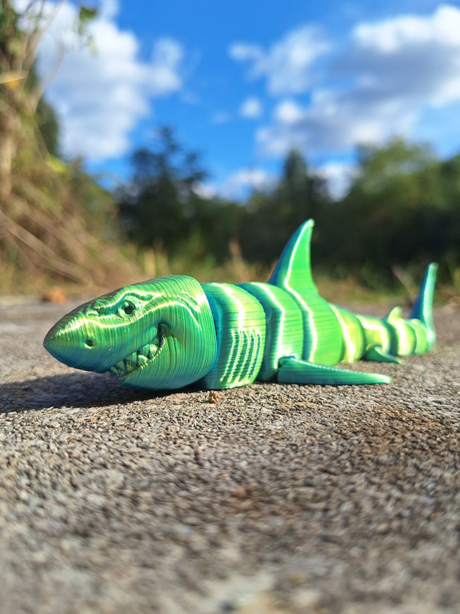 Articulated Great White Shark model printed in Aceaddity Blue/Yellow/Purple tri-color silk PLA filament, showcasing vibrant green and blue stripes, posed on a concrete surface with a natural backdrop of sky and foliage.