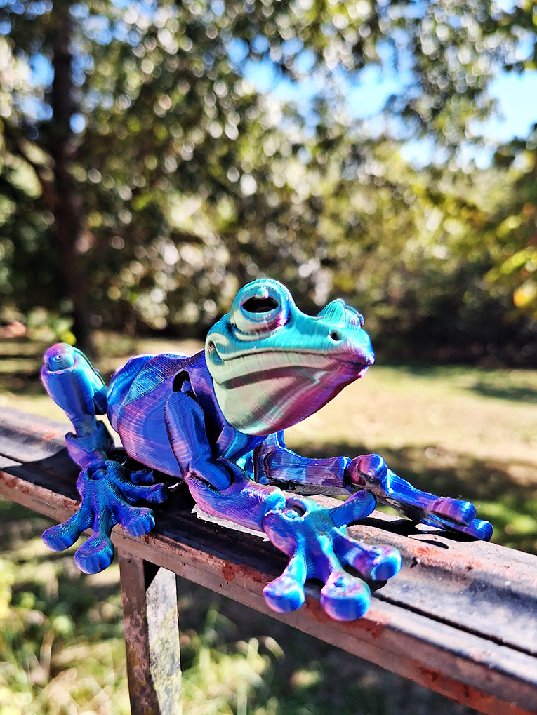 An articulated, magnetic tree frog figure perched on a metal railing, 3D-printed in striking Aceaddity Tri-Color Silk PLA with red, blue, and green tones. The figure's joints are designed to be moved and posed, while the magnets allow it to cling to metal surfaces. The background of lush foliage accentuates the frog's vibrant colors, offering both a decorative and interactive sensory experience.