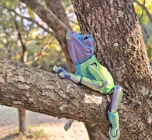 An articulated tree frog figure climbs a tree branch, made from Aceaddity Blue/Yellow/Purple Silk tri-color PLA, which gives it a striking iridescent sheen. The figure's flexible design allows for realistic posing, enhancing its lifelike appearance amidst the tree's rugged bark. The natural outdoor setting highlights the figure's vibrant colors, merging the allure of wildlife with the innovation of 3D printing.