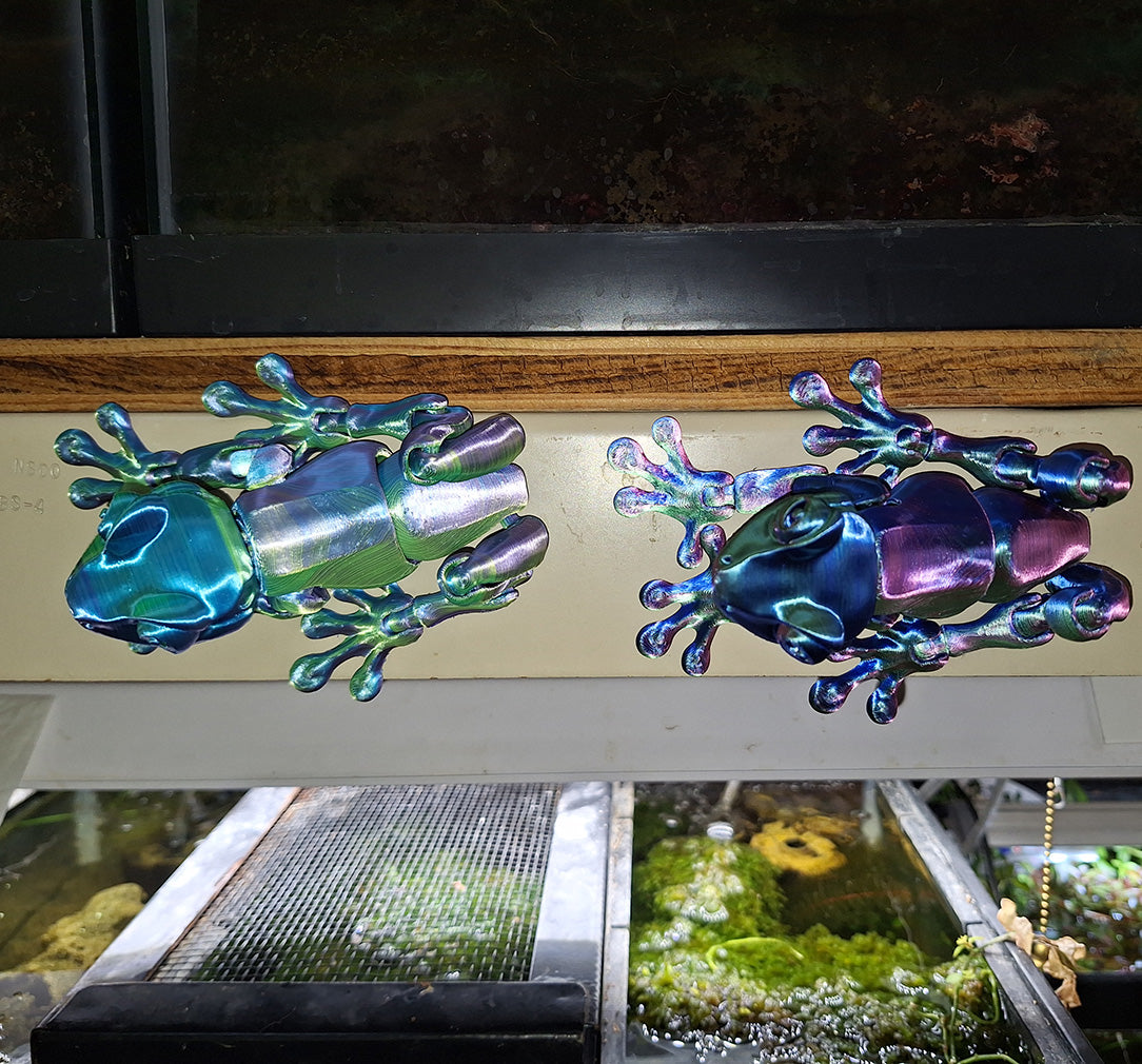 Articulated Tree Frogs with magnetic feet in iridescent Aceaddity tri-color silk PLA, one in hues of blue, yellow, and purple on the left; the other in red, blue, and green on the right, affixed to a metal fish rack, with a vibrant aquarium and foliage in the background.