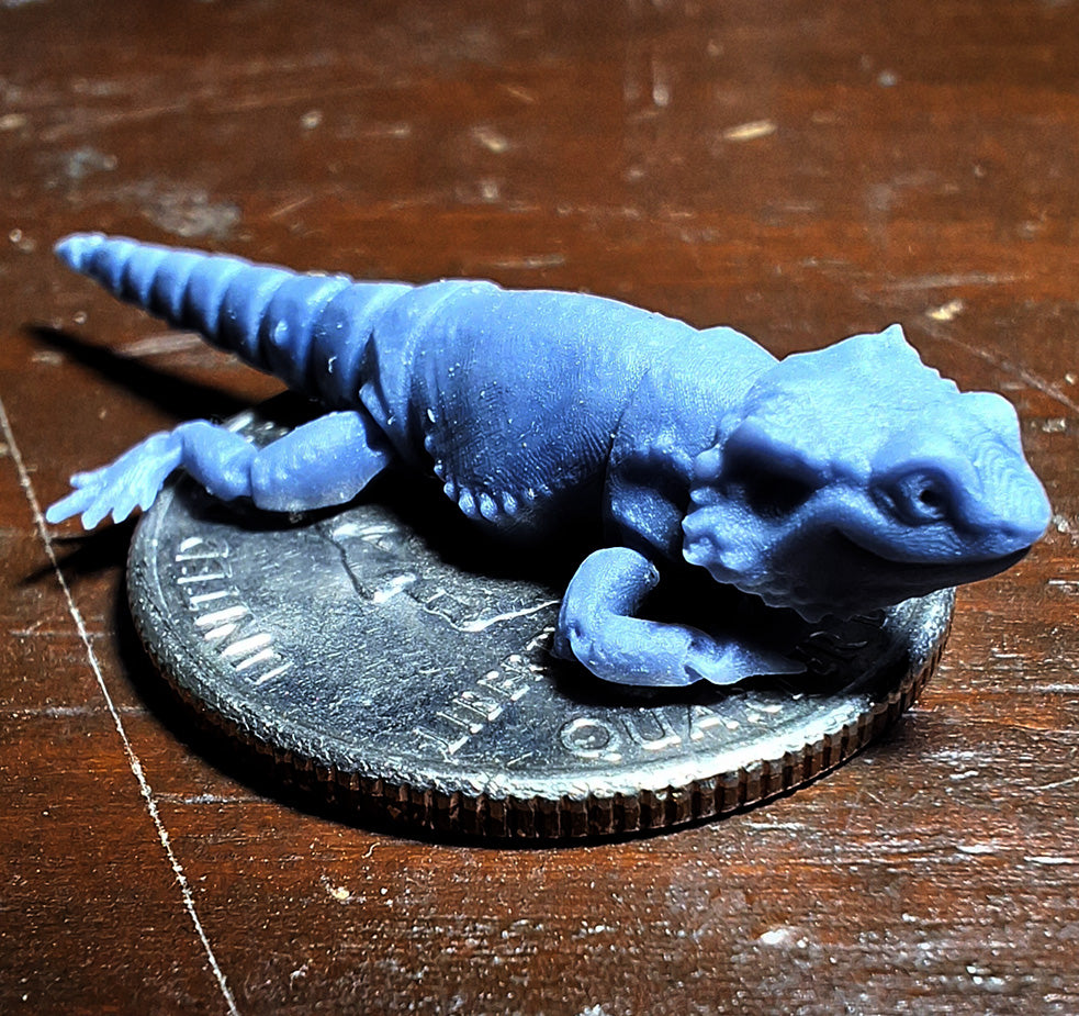 A tiny 3D-printed model of a Bearded Dragon in ABS-like resin, perched atop a quarter to demonstrate its miniature size, from the 'Tinies' line by Cassie's Critters and Creations. Model design by MatMireMakes.