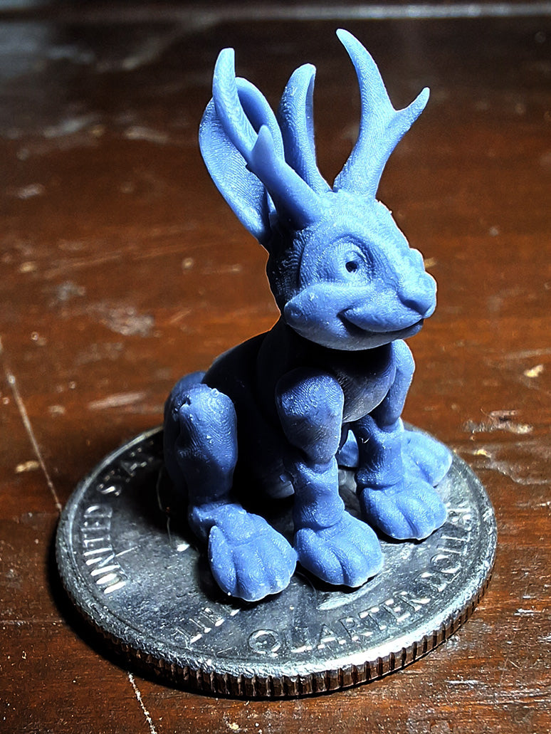 A tiny 3D-printed model of a Jackalope in ABS-like resin, perched atop a quarter to demonstrate its miniature size, from the 'Tinies' line by Cassie's Critters and Creations. Model design by MatMireMakes.
