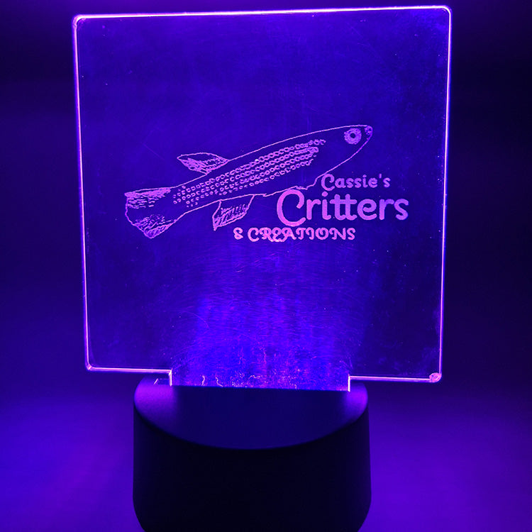 The Cassie's Critters & Creations Logo laser engraved on a square piece of acrylic plastic and mounted in a circular blase base. The base emits a purple glow, illuminating the logo