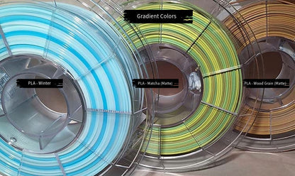 3 spools of filament with gradient colors. From left to right - Winter (white to blue), Matcha (matte yellow to green to brown), and Wood Grain (matte dark brown to tan).