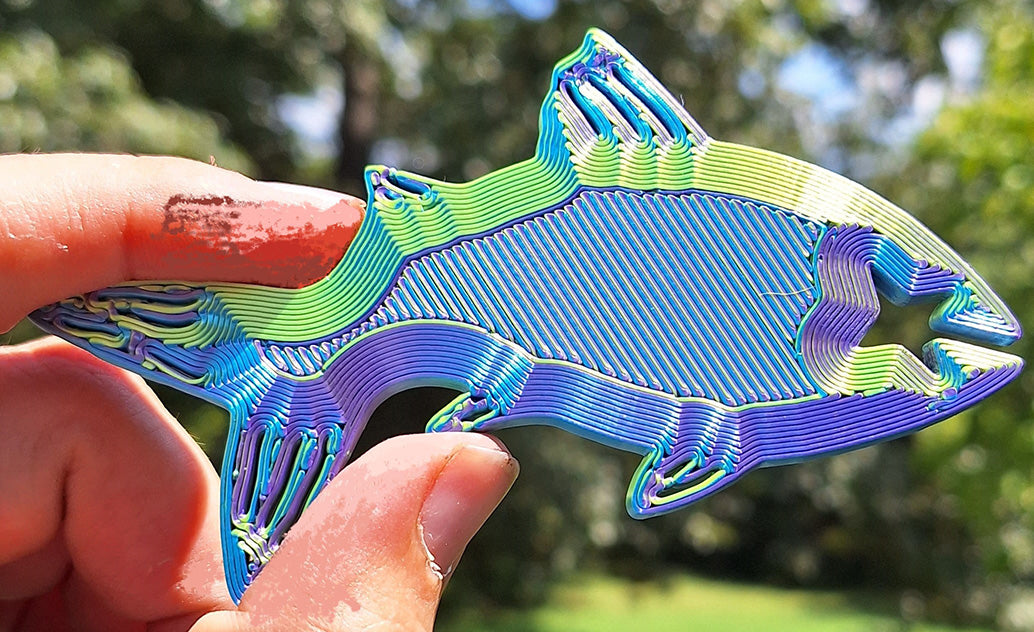 Close-up of a Fishy Bite Bread & Bag clip made with Eryone Red/Yellow/Blue tri-color silk PLA held between fingers, displaying a vibrant color gradient. The clip is pictured in bright outdoor light, showcasing its sleek design and silk-like finish against a natural backdrop.