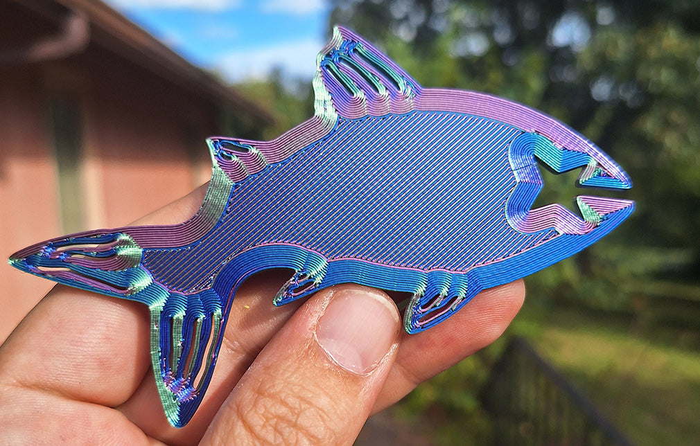 Eryone tri-color silk PLA Fishy Bite Bread & Bag clip in stunning red, blue, and green shades, held up against an outdoor setting with trees and a building in the background. The fish-shaped clip demonstrates its vibrant color shift and glossy finish in natural daylight.