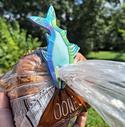 Aceaddity tri-color silk PLA Fishy Bite Bread & Bag clip in vibrant blue, yellow, and purple hues, clamping a bag of Nature's Own whole wheat bread. The clip, shaped like a fish, is showcased outdoors with a natural green backdrop, illustrating its practical use and colorful design.