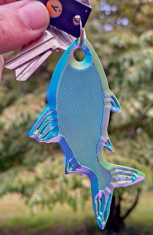 A set of keys attached to a colorful Eryone Red/Yellow/Blue tri-color silk PLA Fishy Bite Keychain, held aloft with a natural, blurred greenery background. The keychain's iridescent colors shine brightly, offering both practicality and a pop of color to everyday life.