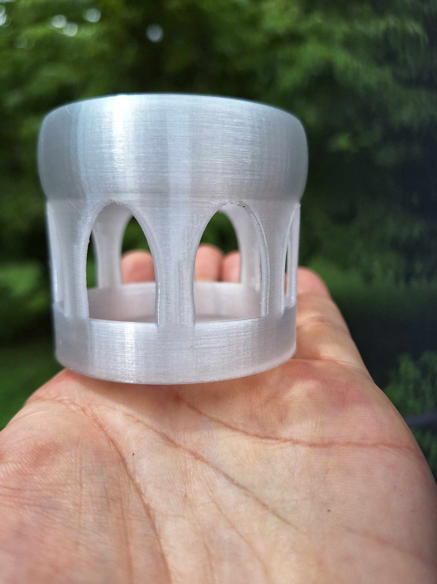 A hand holding a translucent Floating Shrimp Gazebo Feeder by Cassie's Critters & Creations, 3D-printed in Overture Translucent PETG, showcasing its sleek design and clarity against a natural outdoor backdrop.