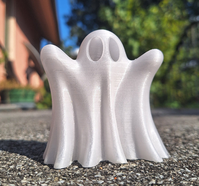 A 'Ghost Booh' figurine 3D-printed in Giantarm glow-in-the-dark purple PLA showcased in white, standing on a concrete surface with a bright blue sky and lush trees in the background, highlighting its playful and spooky design.