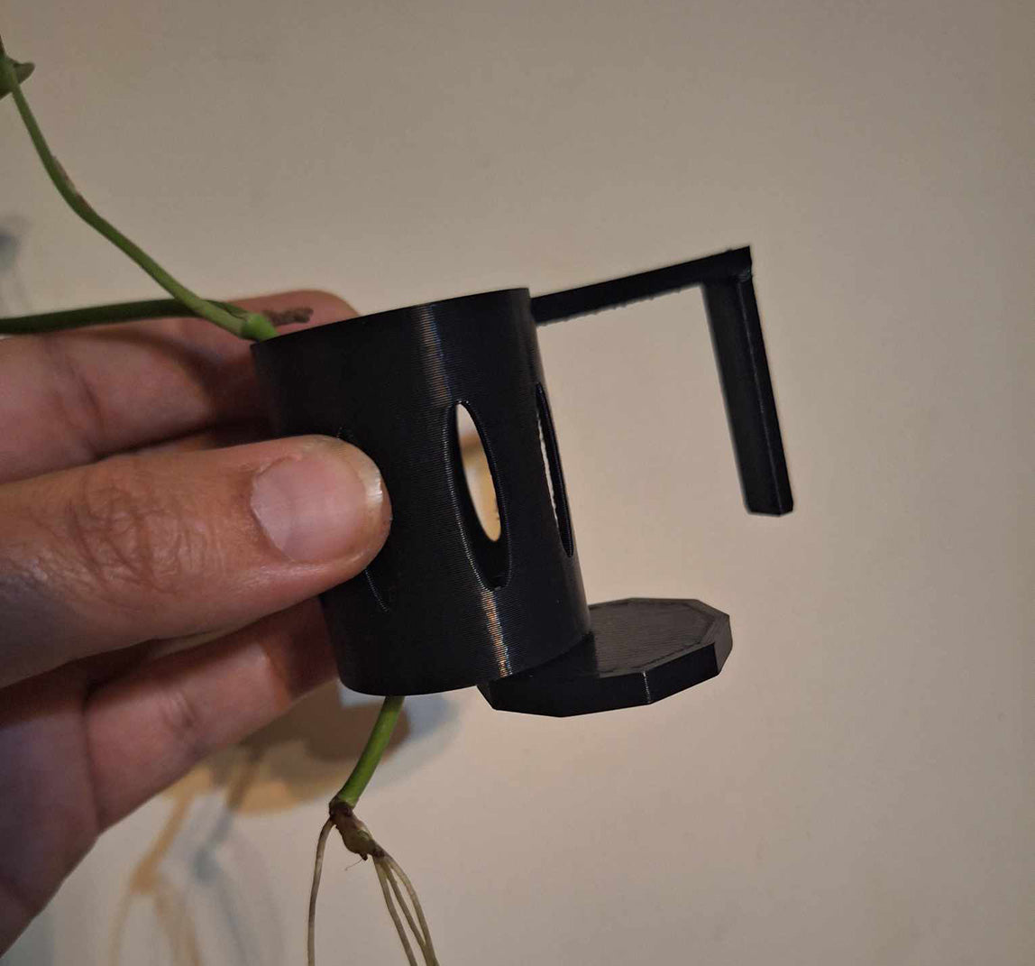 A hand holding a black Hang-On Plant Buddy rimmed aquarium tanks, featuring a built-in leveler. The accessory is 3D-printed with Overture PETG filament and shown with a plant's roots passing through it, illustrating its practical use in supporting aquatic plants.