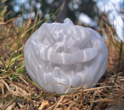 A Jack O' Lantern refrigerator magnet 3D-printed in Giantarm glow-in-the-dark purple PLA, appearing white in daylight, positioned on a forest floor with a backdrop of trees, highlighting its intricate carving details and translucent material.