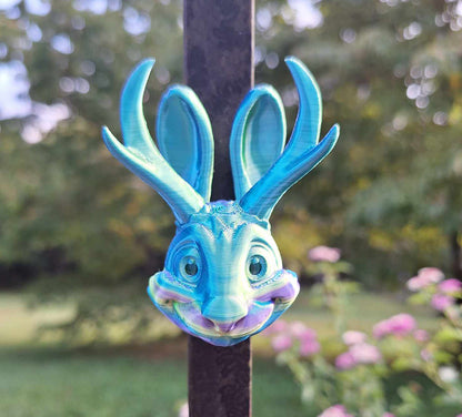 Alt text: "A Jackalope magnet 3D-printed in Aceaddity Blue/Yellow/Purple tri-color silk PLA filament, affixed to a metal pole with a garden setting in the background. The mythical creature magnet features prominent antlers and a playful smile, with iridescent colors that catch the light, perfect for mythical creature collectors and decorative magnet enthusiasts.