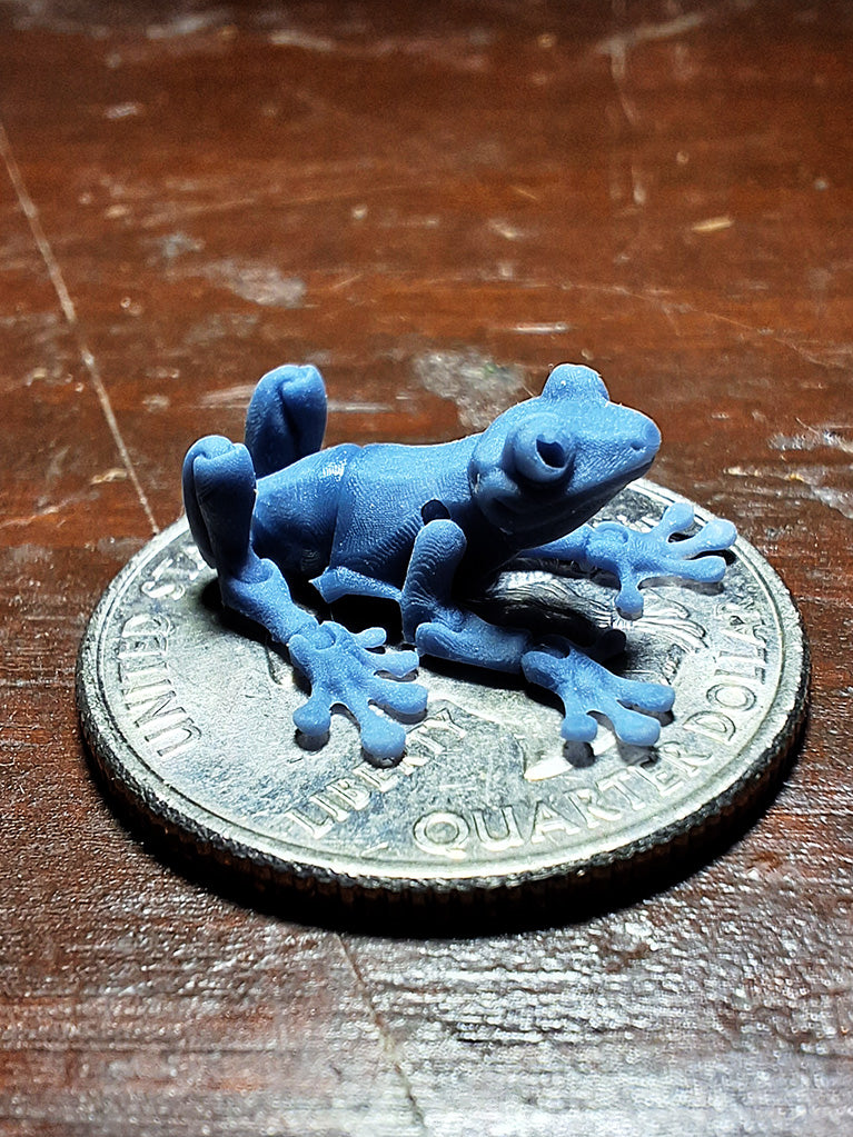 A tiny 3D-printed model of an Articulated Tree Frog in ABS-like resin, perched atop a quarter to demonstrate its miniature size, from the 'Tinies' line by Cassie's Critters and Creations. Model design by MatMireMakes.