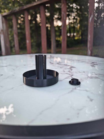 Overture Black PETG Snail Saver Trap displayed on a marble surface, with the cap placed off to the side. The trap's dark color contrasts sharply with the light background, set against an outdoor setting with trees, highlighting its sleek design and functional components.