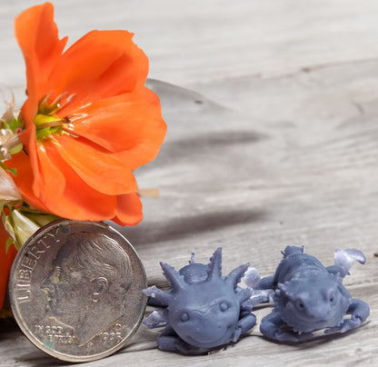 Miniature 3D-printed axolotl and bearded dragon models beside a US dime for scale, with a vibrant orange flower accent, all against a rustic wood grain background.