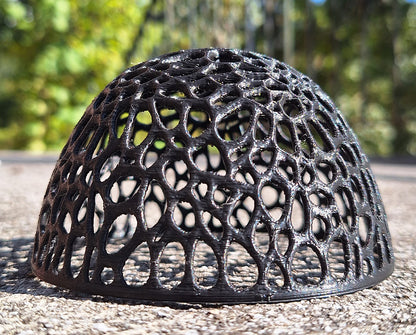 The VoroDome Shrimp Sanctuary, a voronoi tessellation sculpture, sits on a concrete surface. It's 3D-printed in Creality Hyper Black PLA, creating a striking contrast with its environment. The dome's openwork design casts delicate shadows, offering a unique aesthetic that blends art with functionality in an outdoor setting.